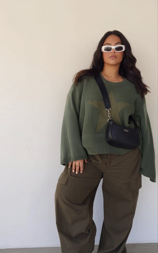 Woman in an oversized olive green star knit sweater and cargo pants posing against a wall.