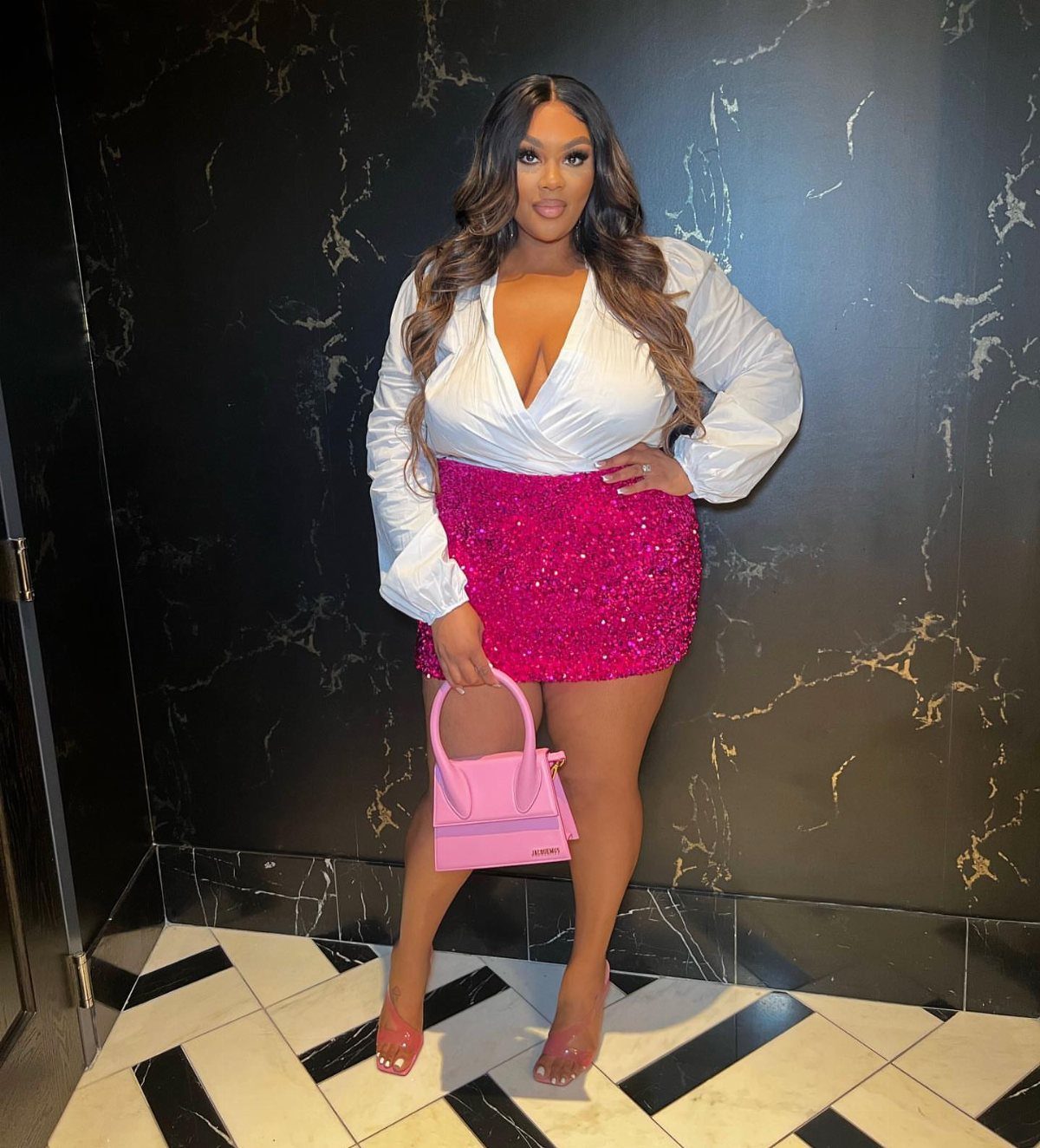 Woman posing in a white wrap top and pink sequined mini skirt holding a pink handbag.