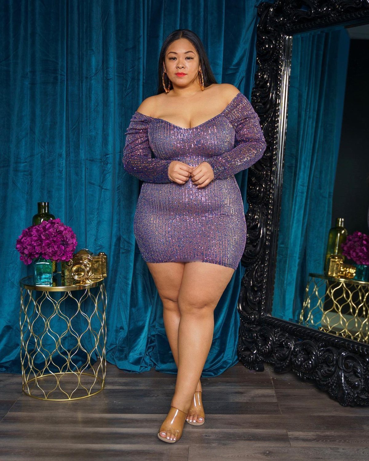 A woman wearing a sparkling purple off-the-shoulder mini dress posing against teal curtains.