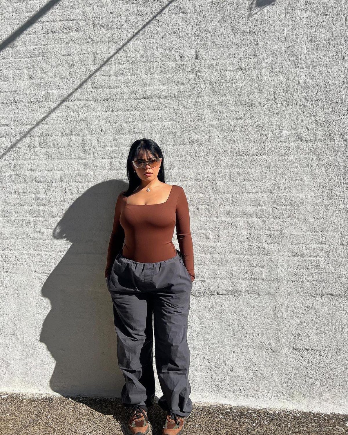 A woman in a brown bodysuit and grey trousers standing against a white brick wall.