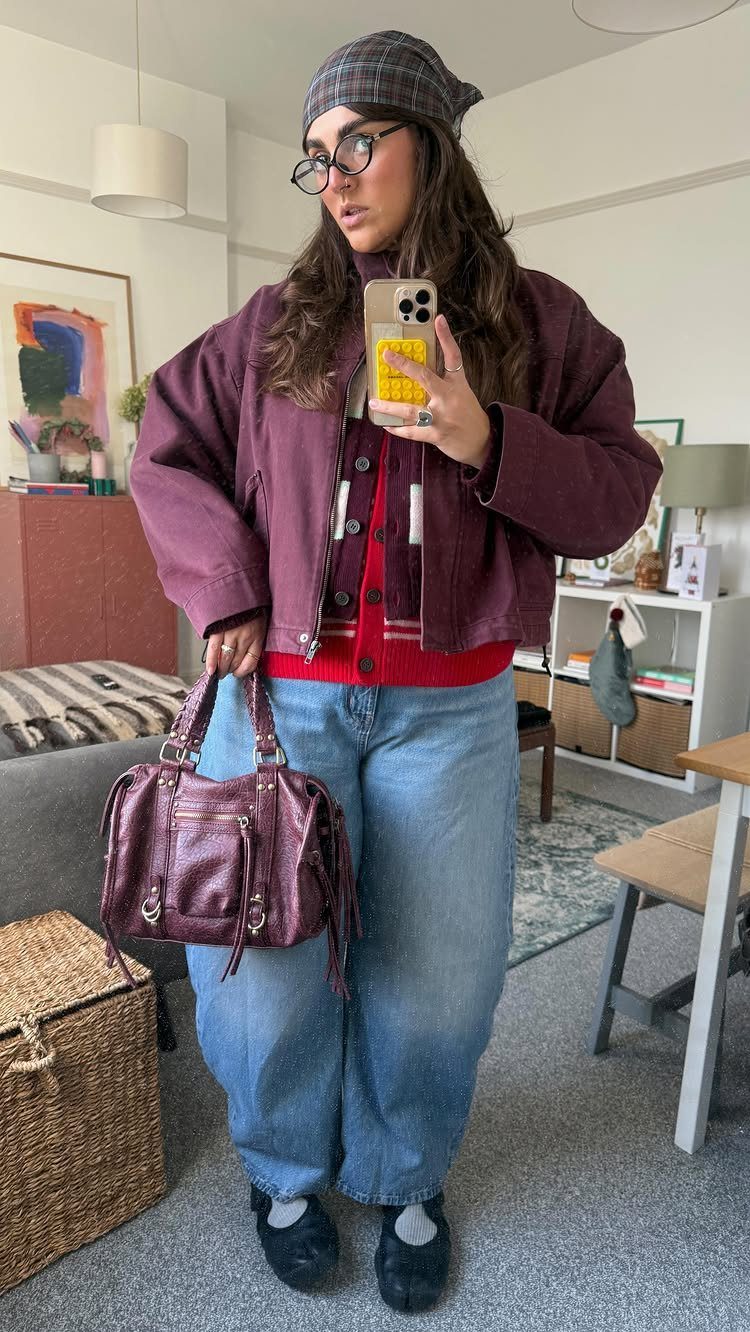 Woman in a maroon jacket and red cardigan layered over blue jeans and holding a handbag.