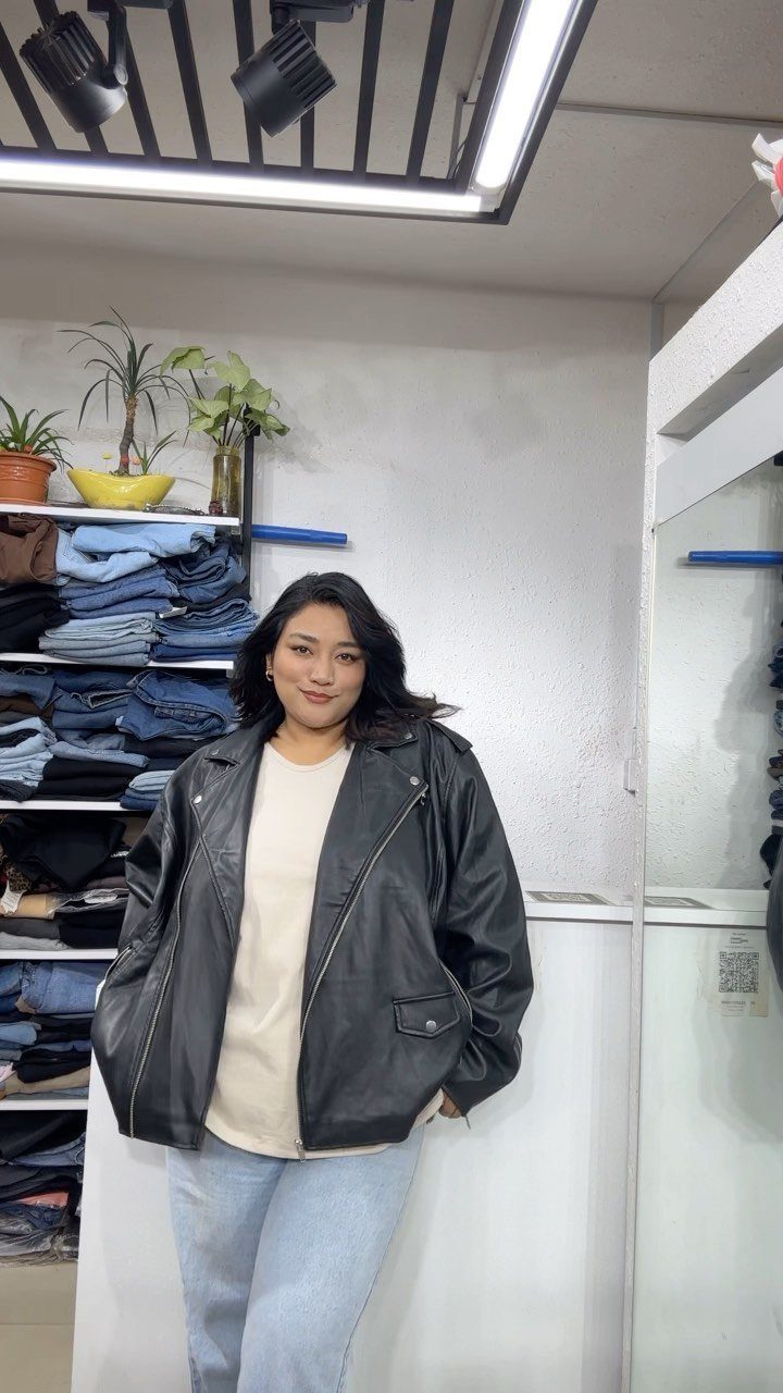 A woman posing in a black leather jacket, cream shirt, and light blue jeans.