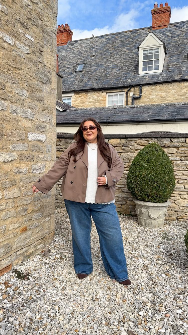 A woman wearing a tan unstructured trench coat and wide-leg blue jeans standing outdoors.