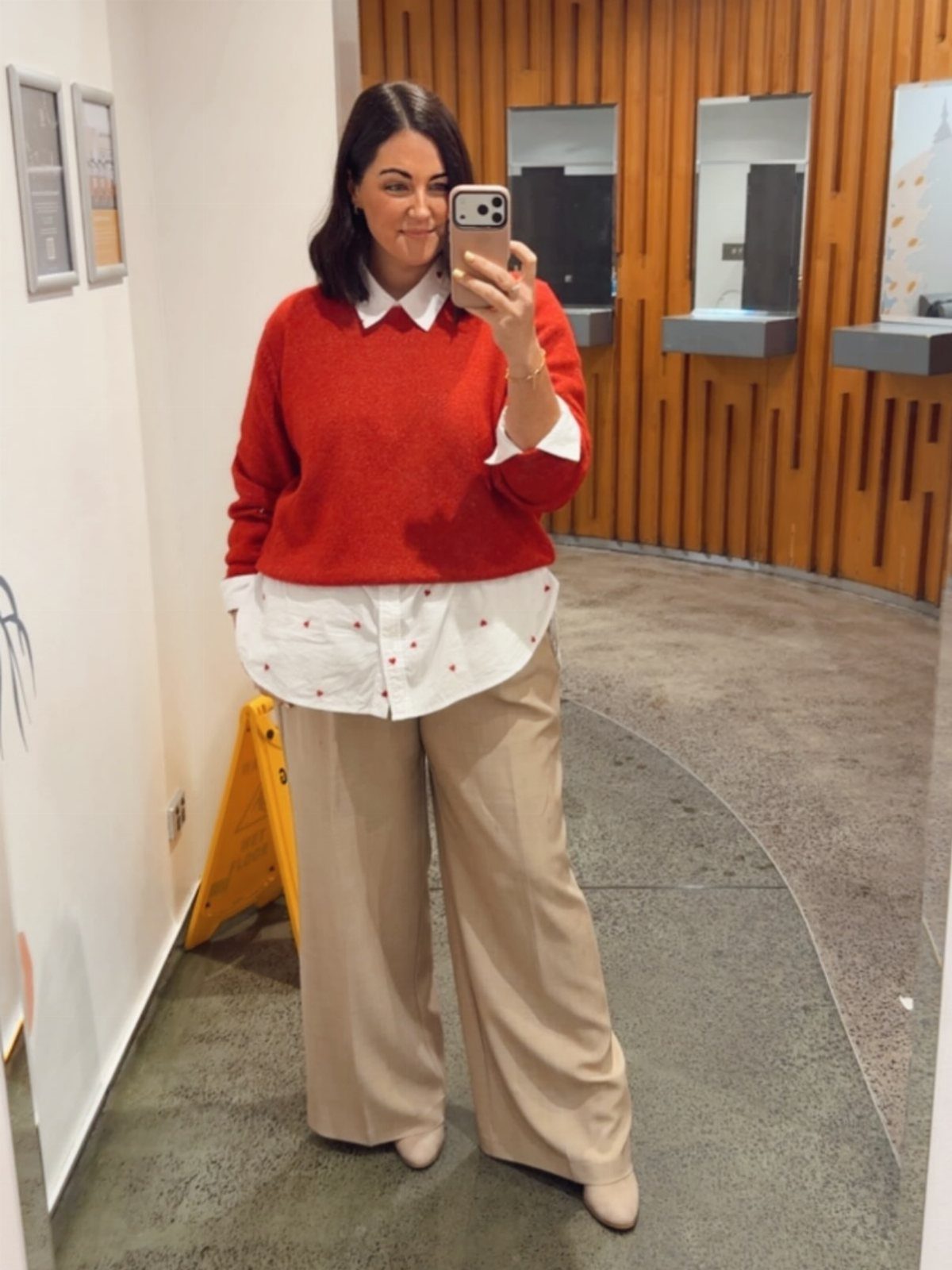 A woman posing in a red sweater, white collared shirt, and beige wide-leg trousers.