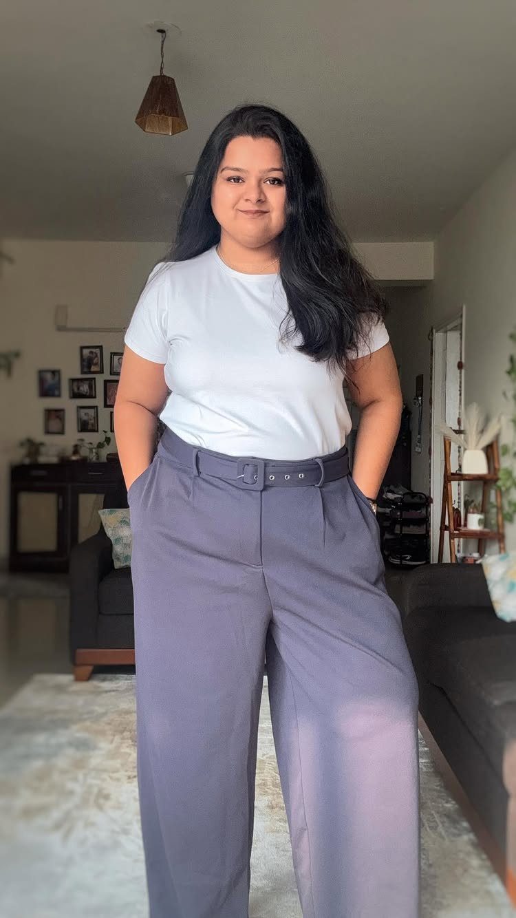 A smiling woman wearing a white t-shirt and wide-leg purple trousers standing in a living room.