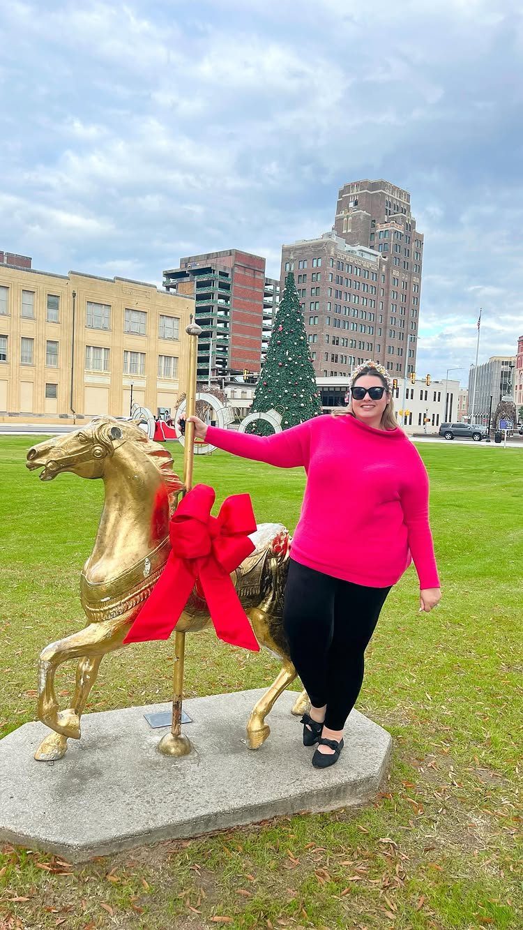 Woman in a pink sweater and black leggings standing by a gold carousel horse.