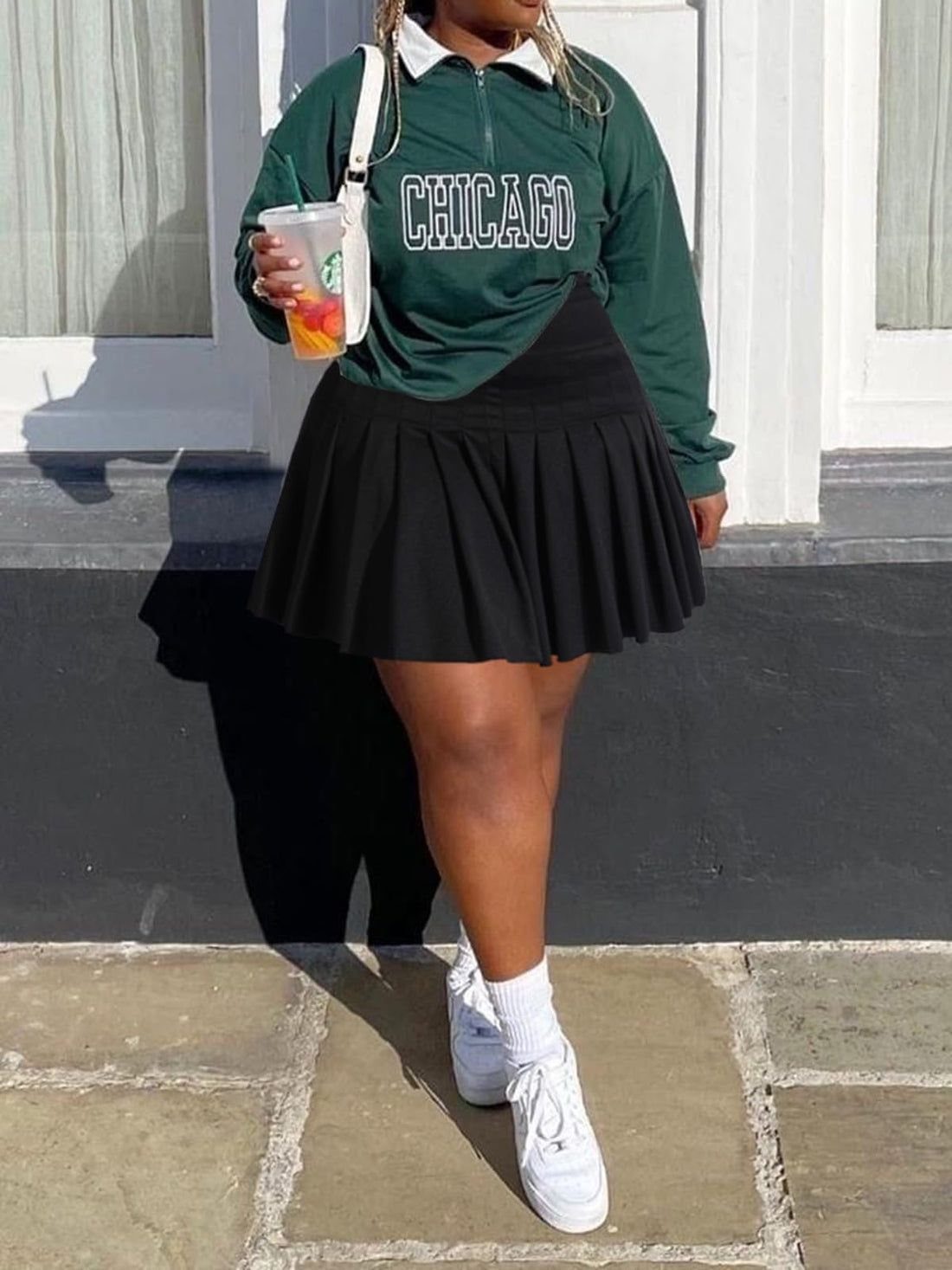 A person wearing a green Chicago sweatshirt and a black pleated skater skirt with white sneakers.