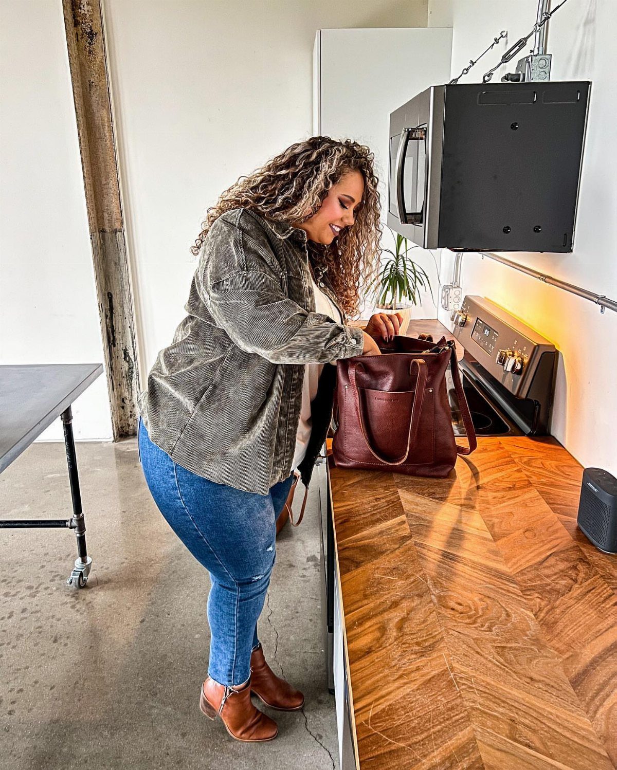 A woman in a patterned shirt and blue jeans looks into a brown leather tote bag on a wooden counter.