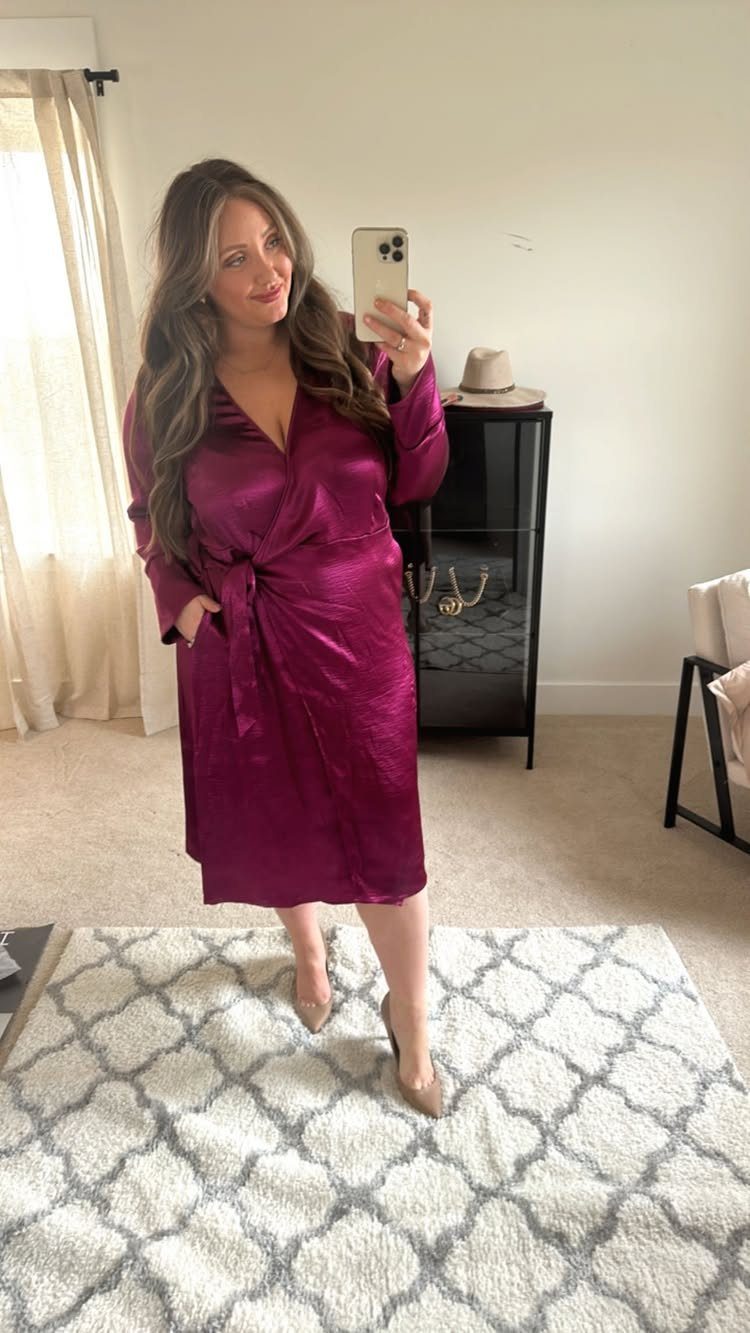 Woman in a burgundy satin wrap dress posing for a mirror selfie indoors.