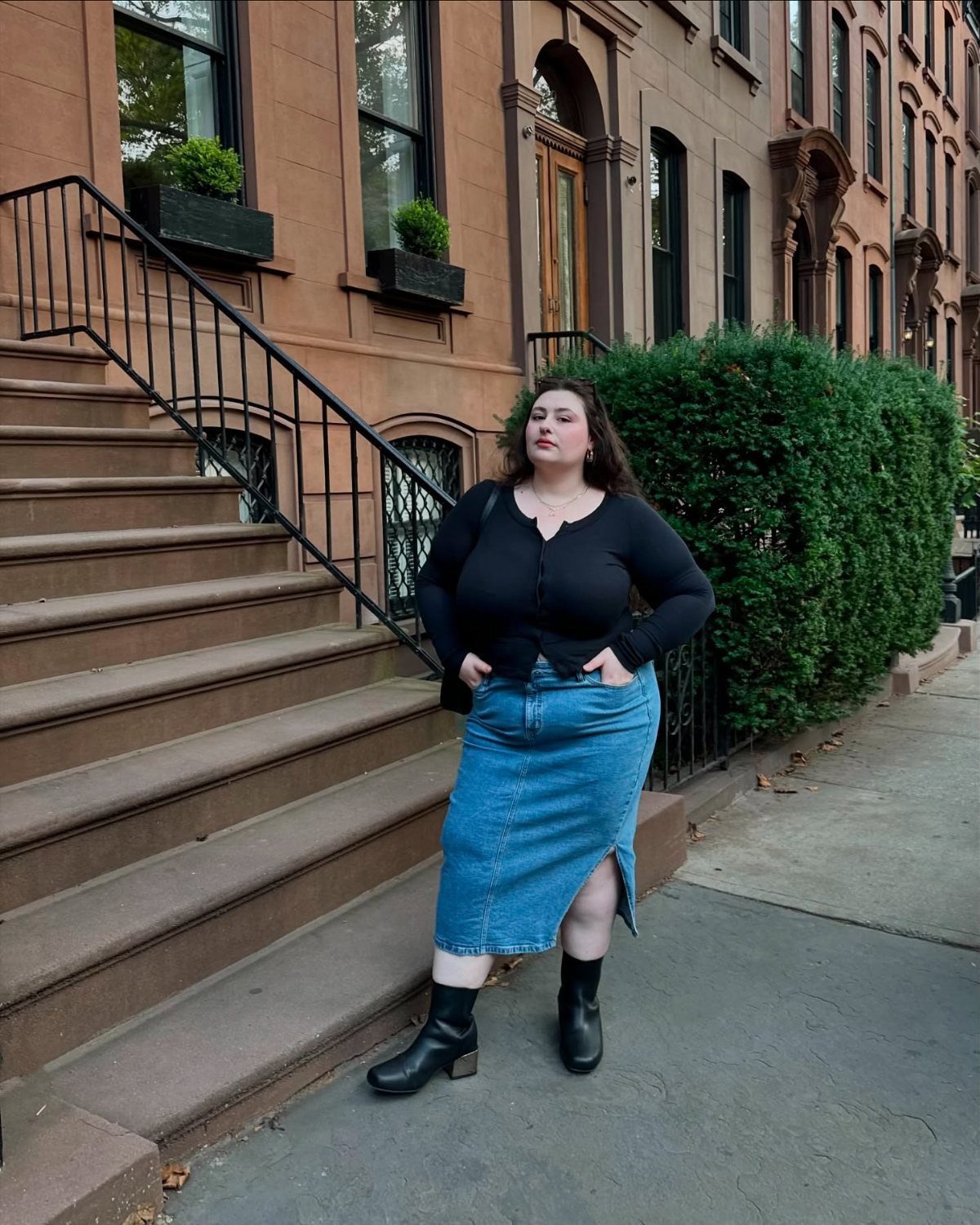 A woman in a black top and denim skirt posing on city steps wearing black ankle boots.