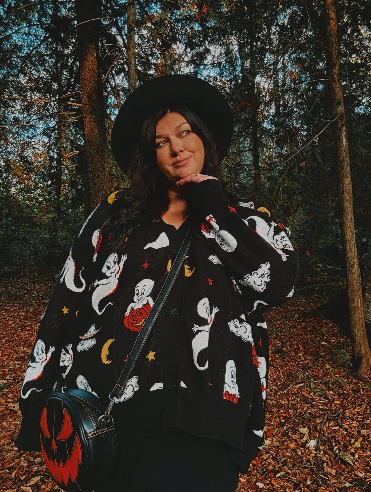 Woman in a black ghost and pumpkin print cardigan posing in a forest setting.