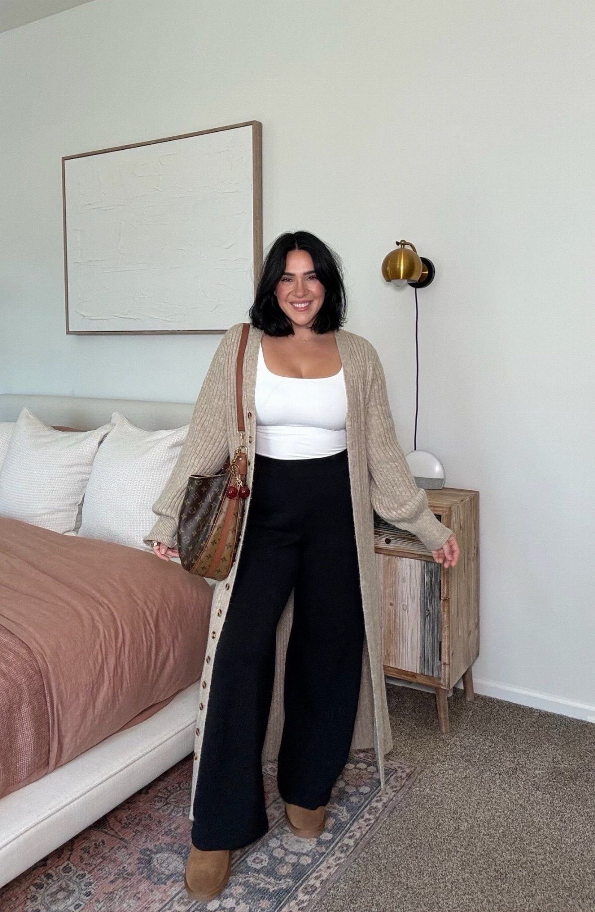 A smiling woman wearing a long beige knit cardigan, white top, and black wide-leg trousers.