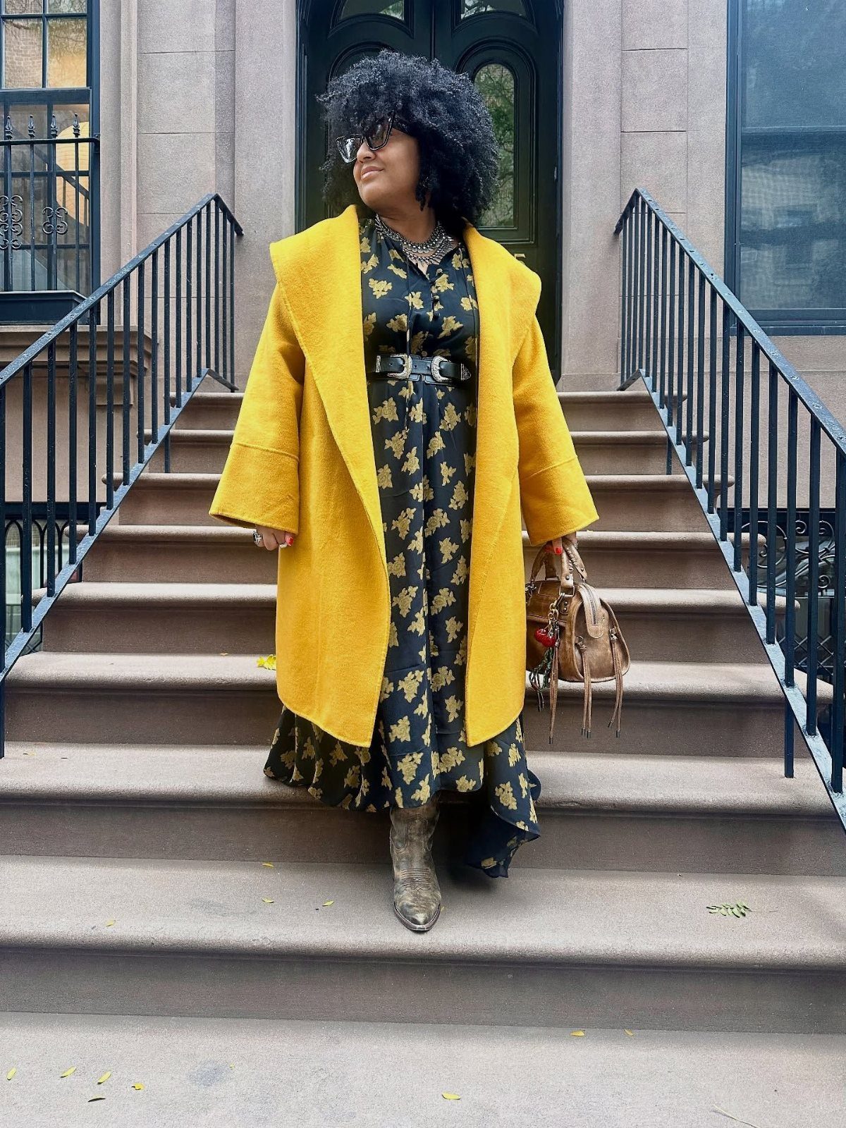 A woman in a mustard yellow wool coat and floral dress standing on outdoor stairs.