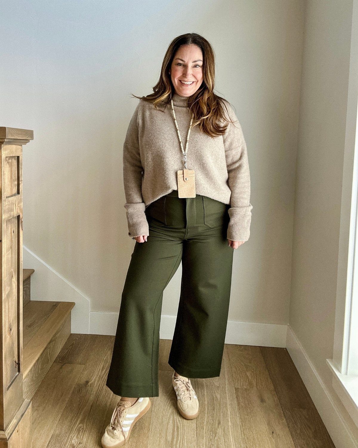 A smiling woman wearing a beige sweater and olive green wide-leg trousers standing indoors.