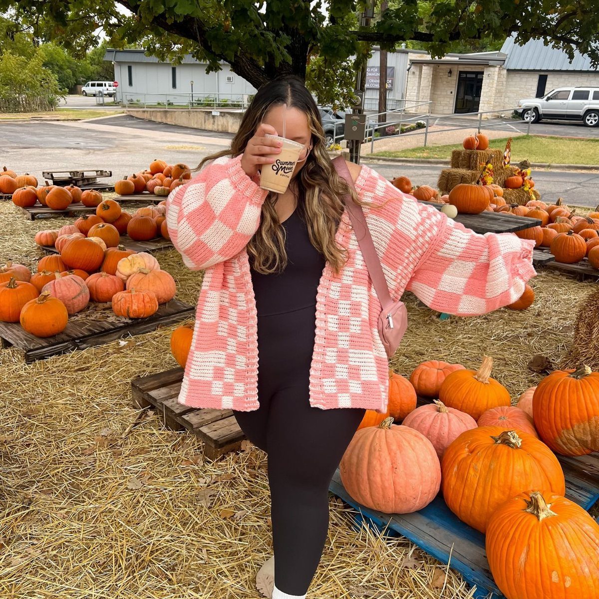 Woman in a pink checkered cardigan standing amidst many orange pumpkins in a field.