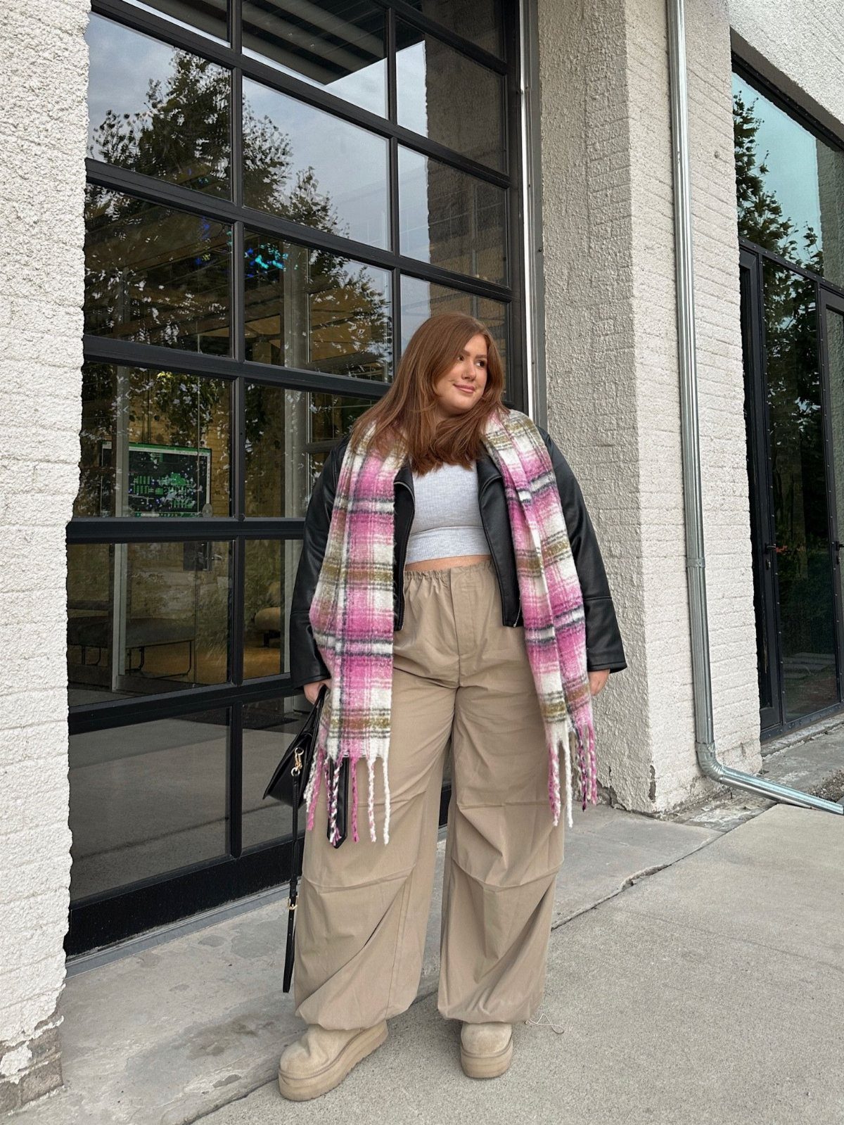 A woman wearing a black leather jacket and a large pink plaid scarf standing outdoors.