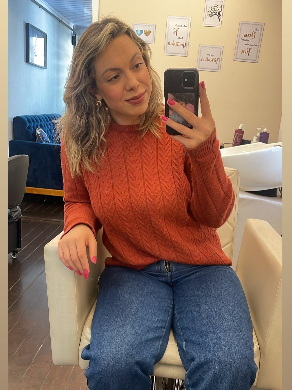 A woman wearing a rust orange cable-knit sweater and blue jeans sitting in a chair.