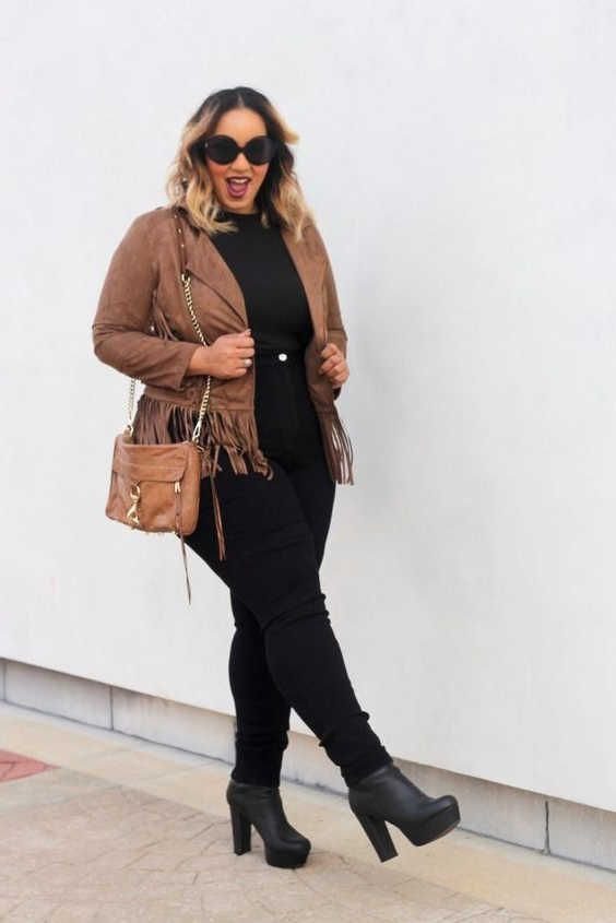 Woman in a tan suede fringe jacket, black outfit, and platform boots posing against a white wall.