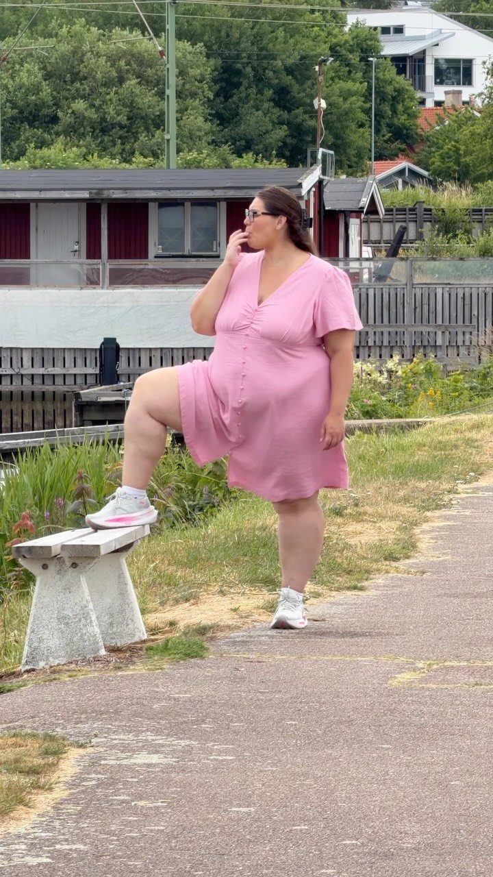 A woman in a pink summer dress and white sneakers posing outdoors near a bench.