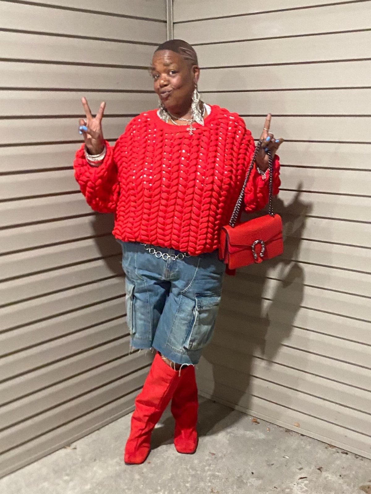 A person wearing a red chunky knit sweater, denim shorts, and red boots posing against a wall.