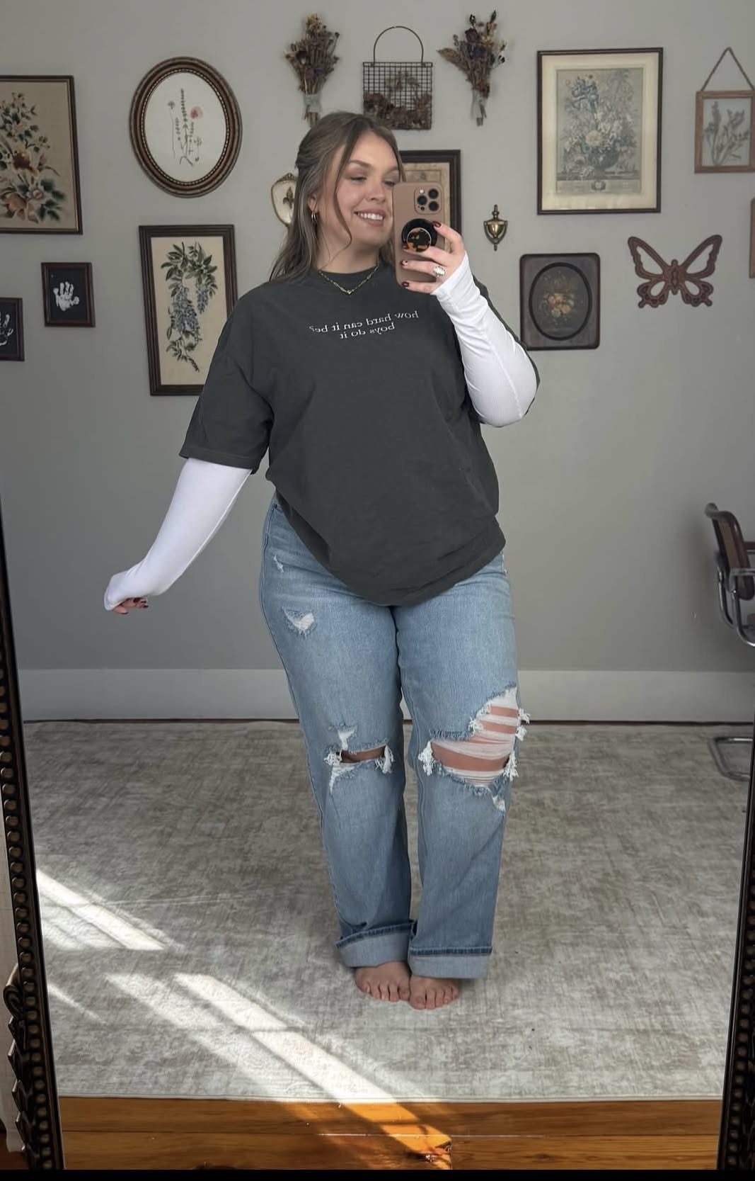 Woman posing in a dark graphic t-shirt and ripped denim jeans for a mirror selfie.