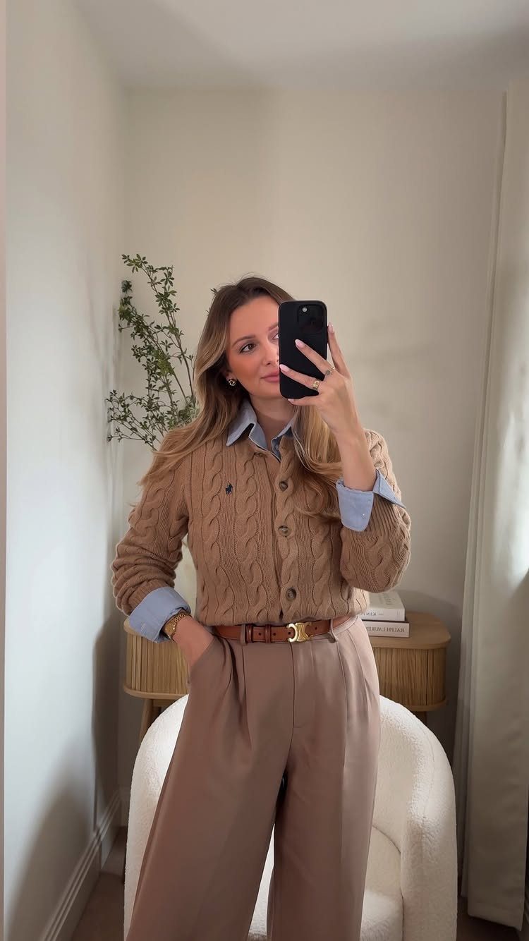 Woman in a tan cable-knit cardigan, blue shirt, and beige trousers taking a mirror selfie.