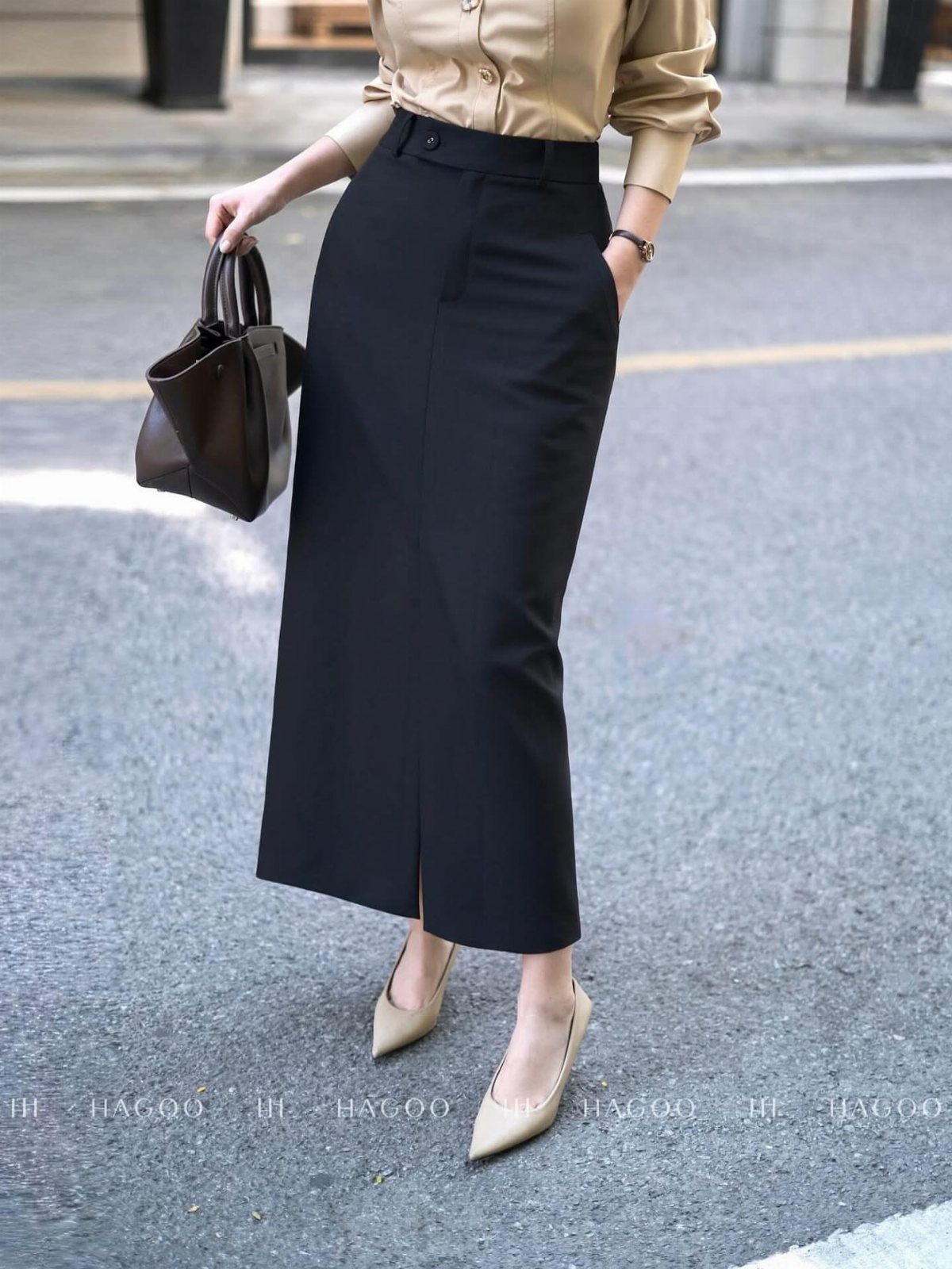 A person wearing a long black midi skirt, beige blouse, and beige heels holding a handbag.