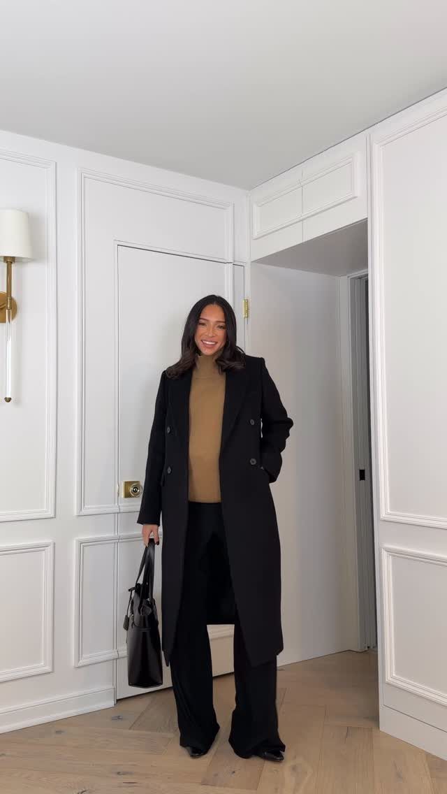 Smiling woman wearing a long black coat, tan turtleneck, and black trousers while holding a handbag.