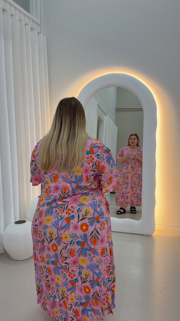 Woman in a pink floral maxi dress looking at her reflection in an arched backlit mirror.