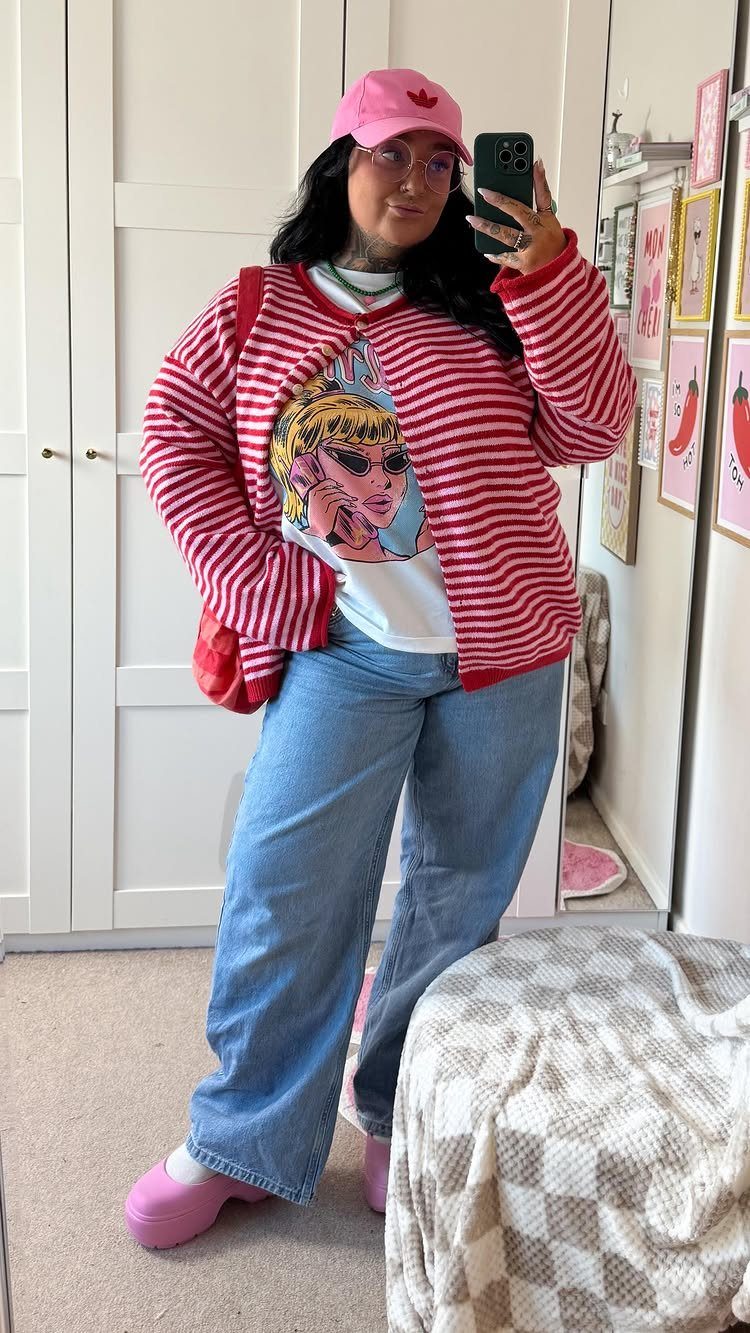 Woman in a graphic tee, striped cardigan, and blue jeans posing for a mirror selfie.