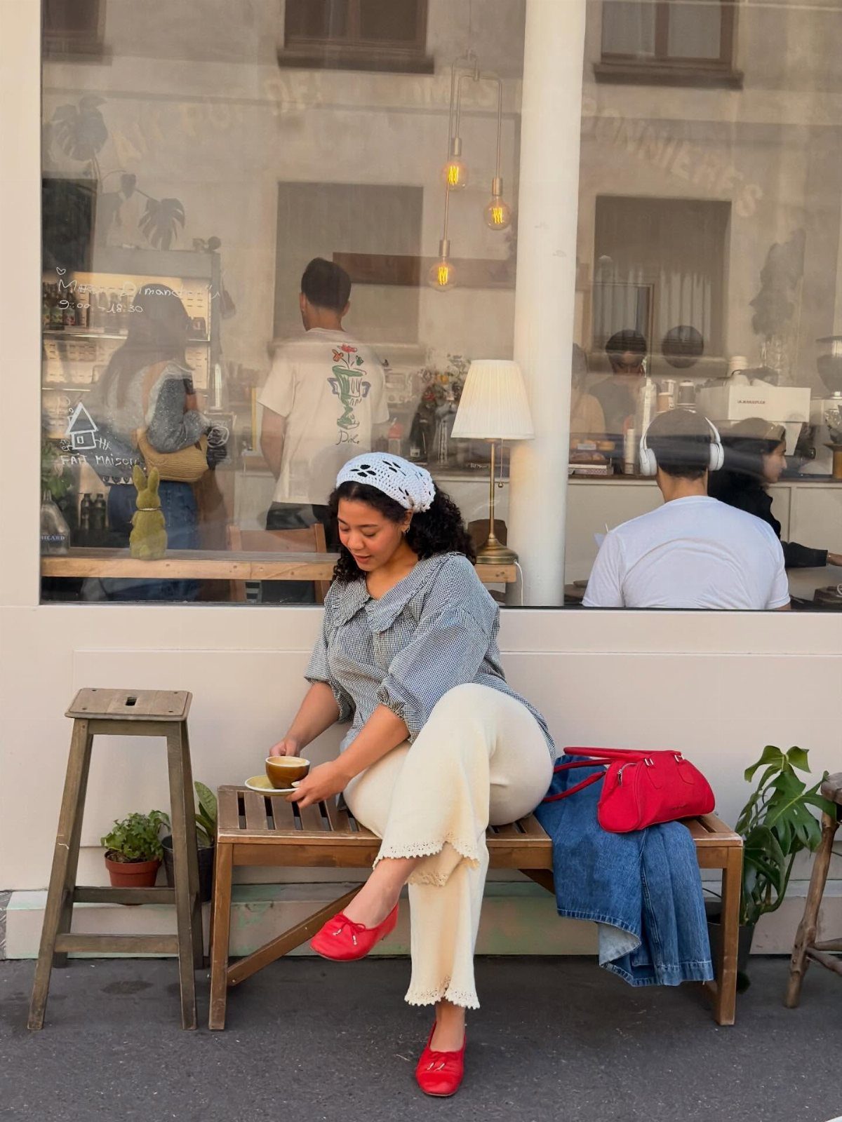 Woman in a patterned blouse and cream trousers sitting on a wooden bench outside a cafe.