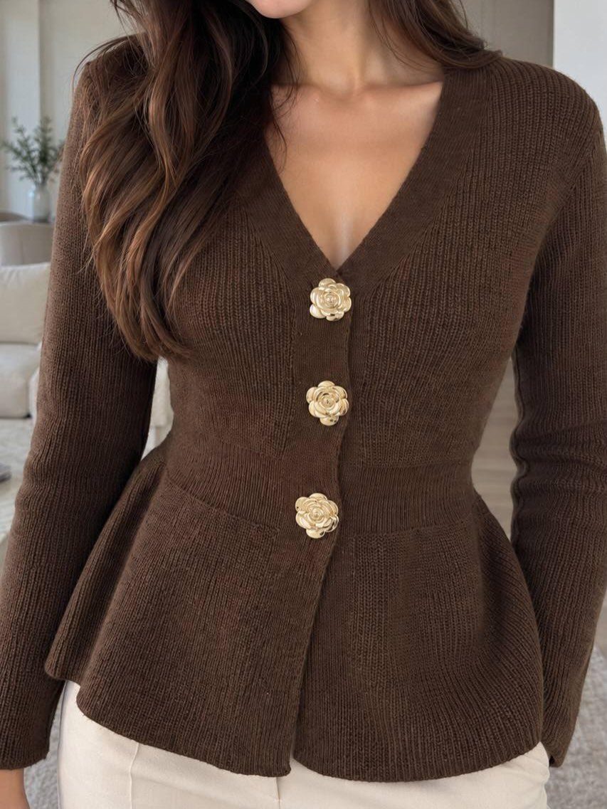 A person wearing a brown ribbed knit cardigan with large gold flower buttons and white trousers.