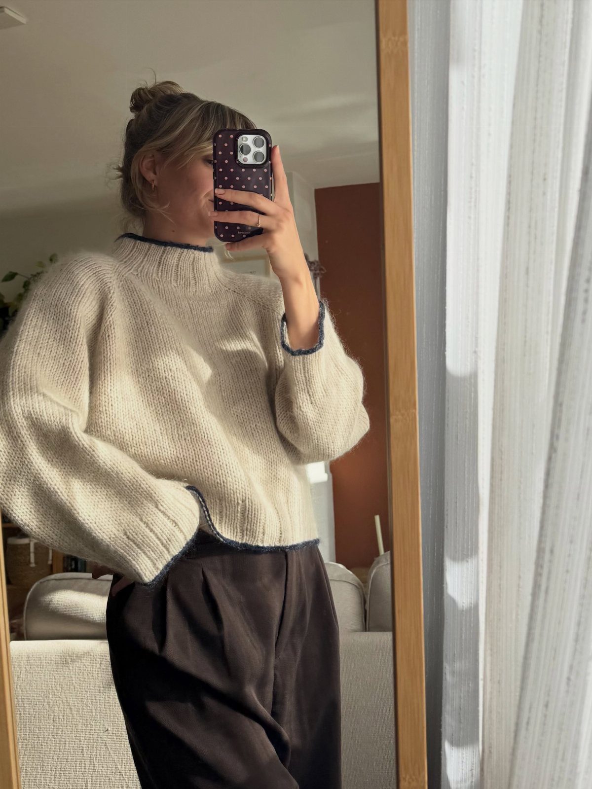 A woman posing in a mirror wearing a cream knit sweater and dark brown trousers.