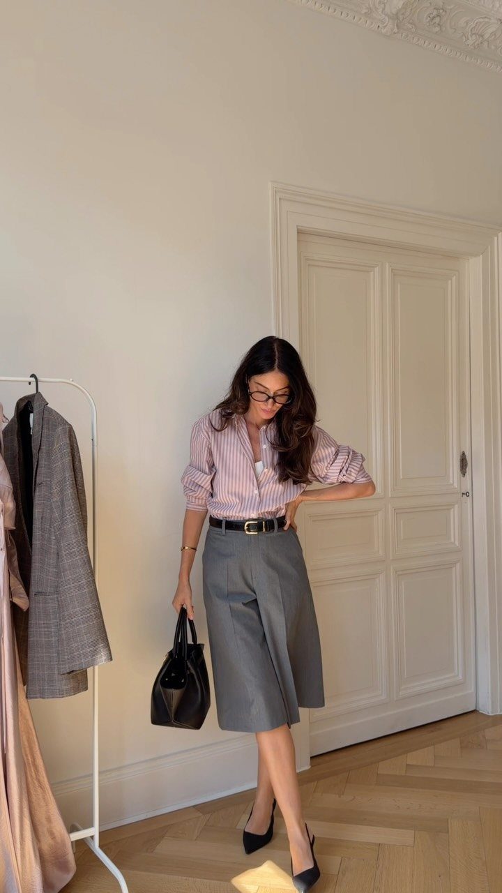 Woman in a pink striped shirt and grey culottes holding a black handbag.