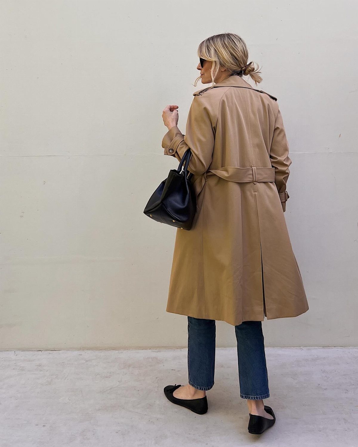 Woman wearing a long khaki trench coat, blue jeans, and black flats against a neutral wall.