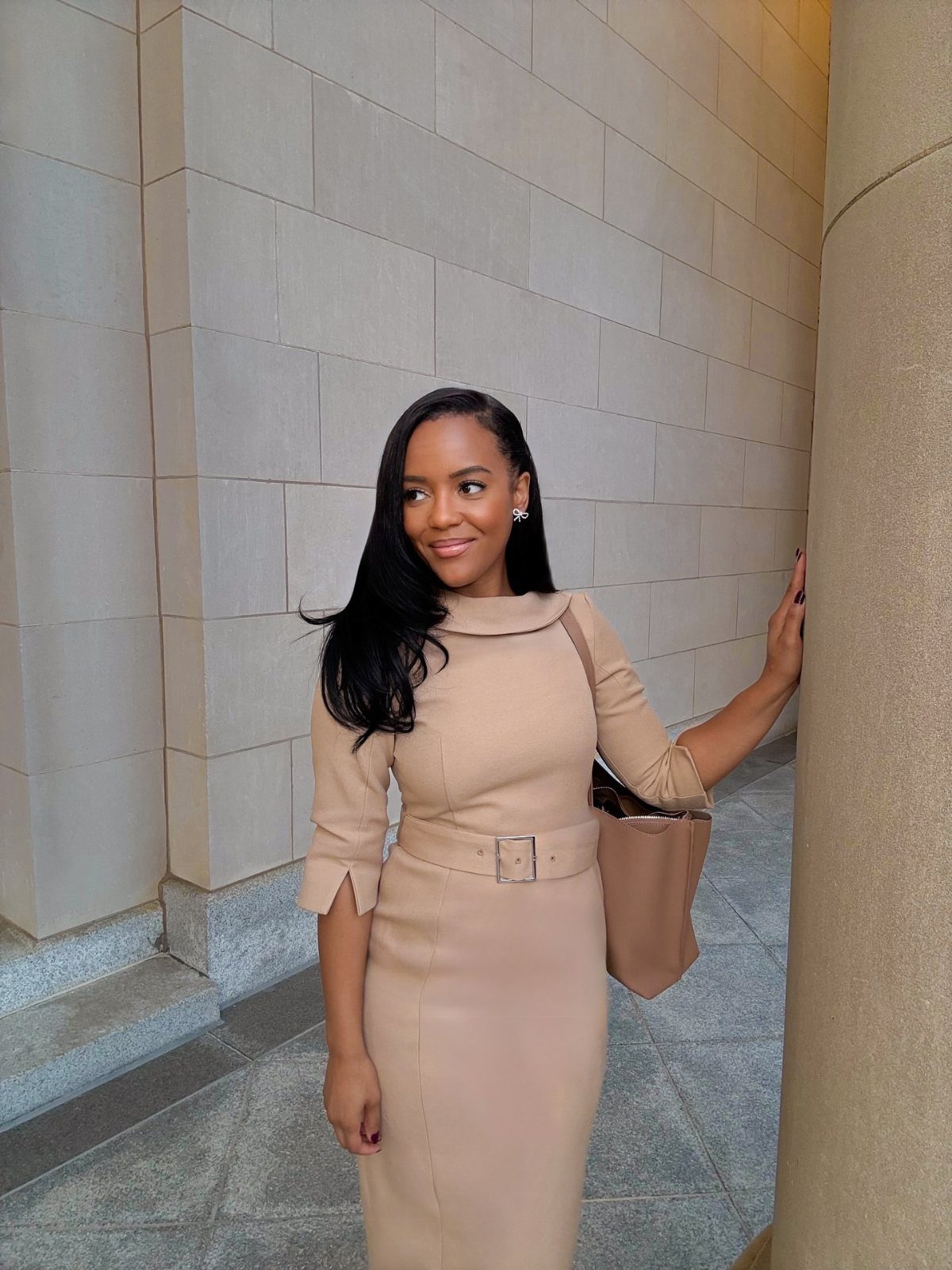A woman wearing a beige long-sleeved midi dress with a matching belt standing by a stone wall.