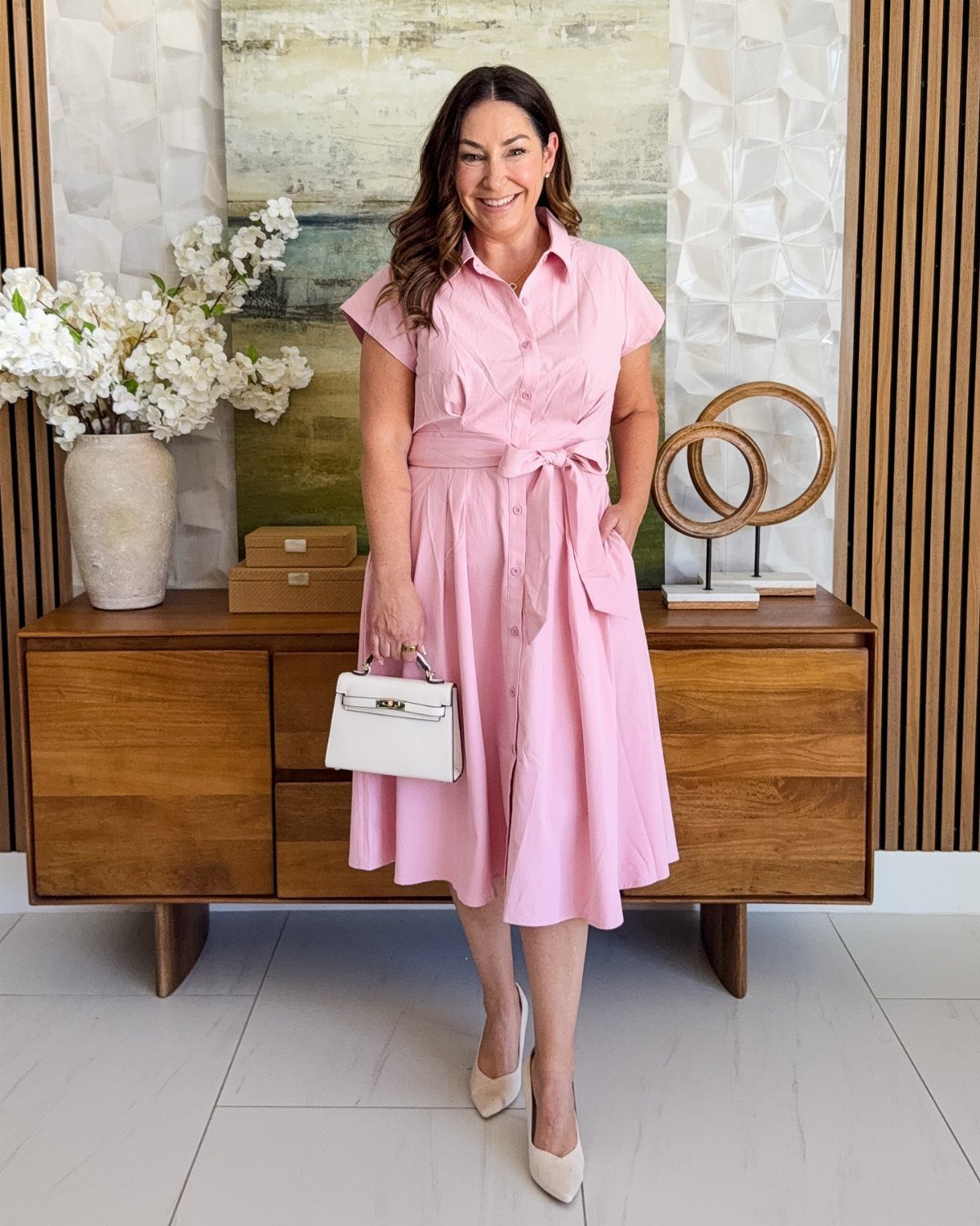 A woman wearing a pink button-down shirt dress and beige pumps standing in a bright room.