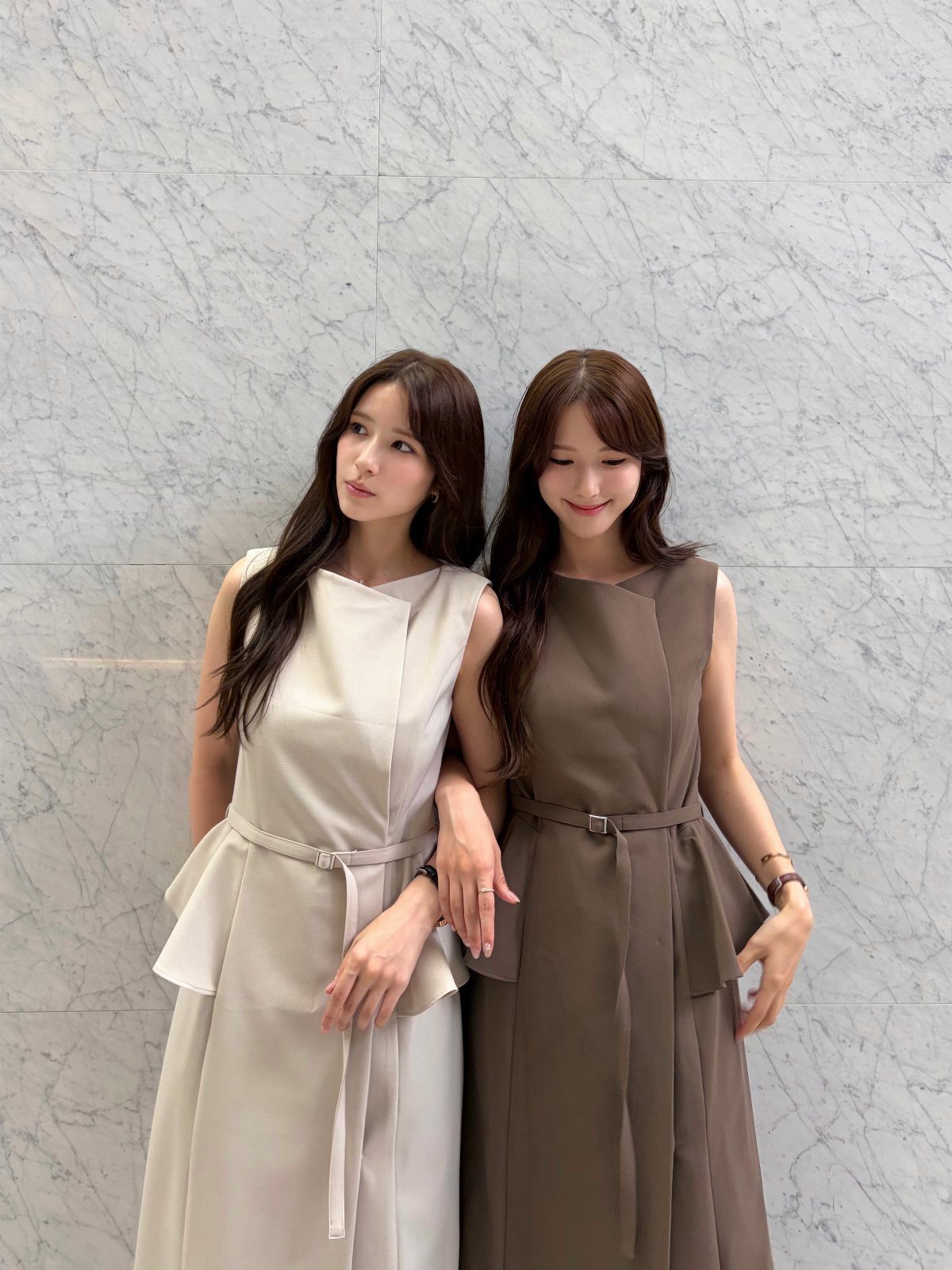 Two women posing in sleeveless peplum dresses against a white marble wall background.