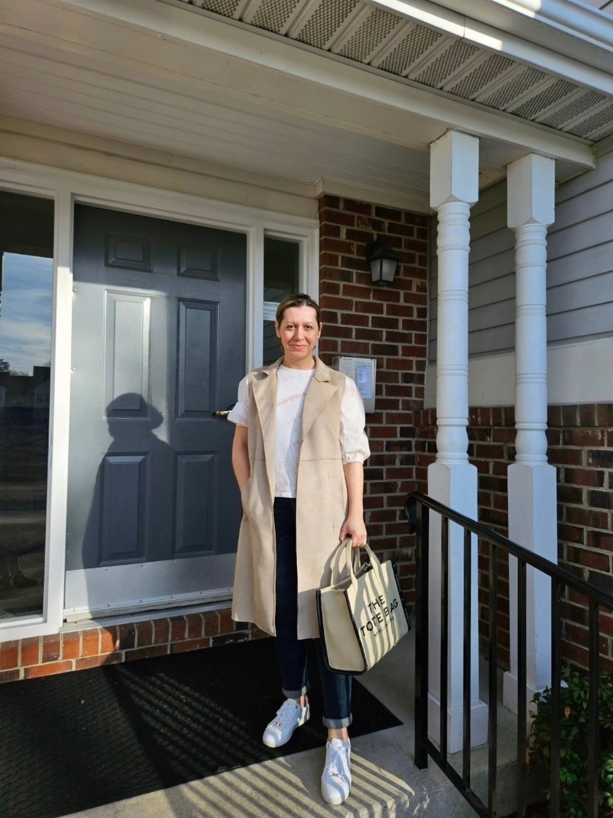 Woman in a long beige vest, white t-shirt, dark denim pants, and white sneakers standing on a porch.