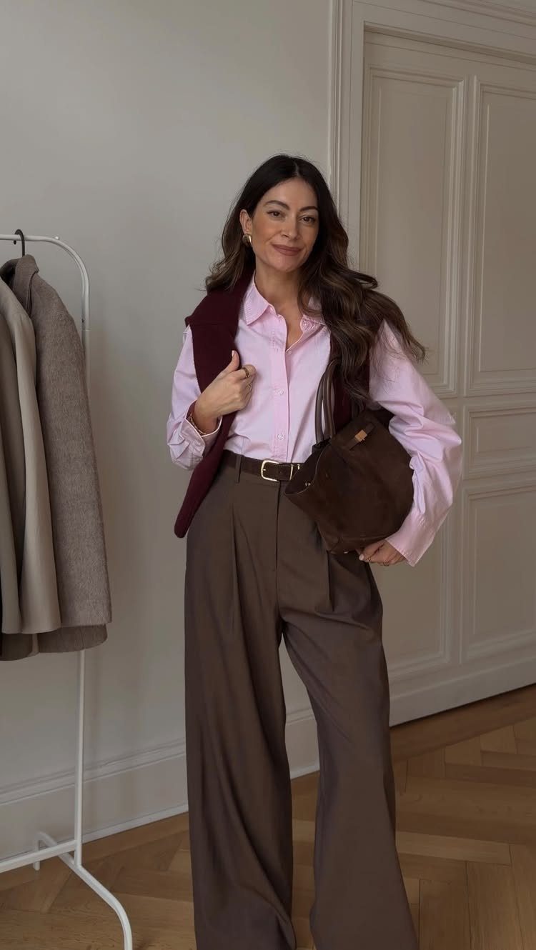 Woman wearing a pink shirt, brown wide-leg trousers, and a burgundy sweater draped over her shoulders.