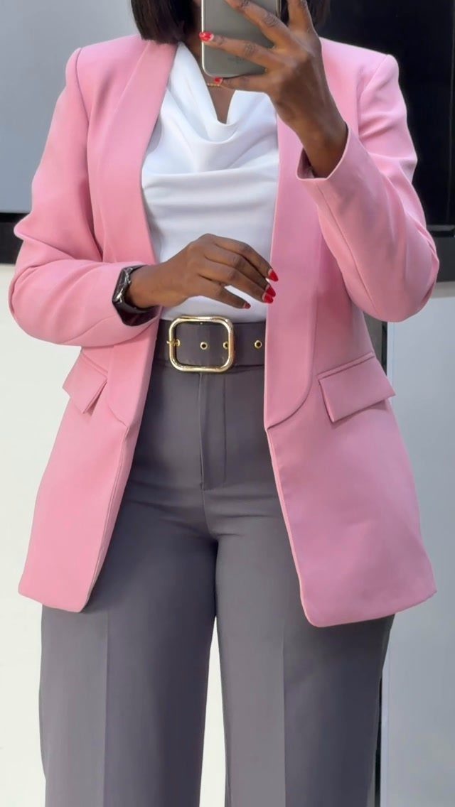 A person wearing a pink blazer, white top, and grey trousers with a gold buckle belt.