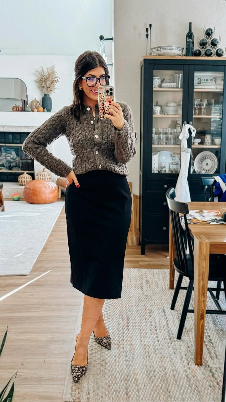 Woman in a brown cardigan and black midi skirt posing for a mirror selfie indoors.