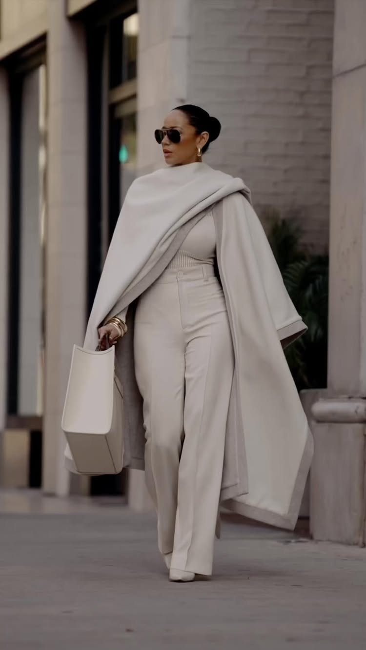 Woman in cream jumpsuit wearing a large draped shawl and carrying a white tote bag.
