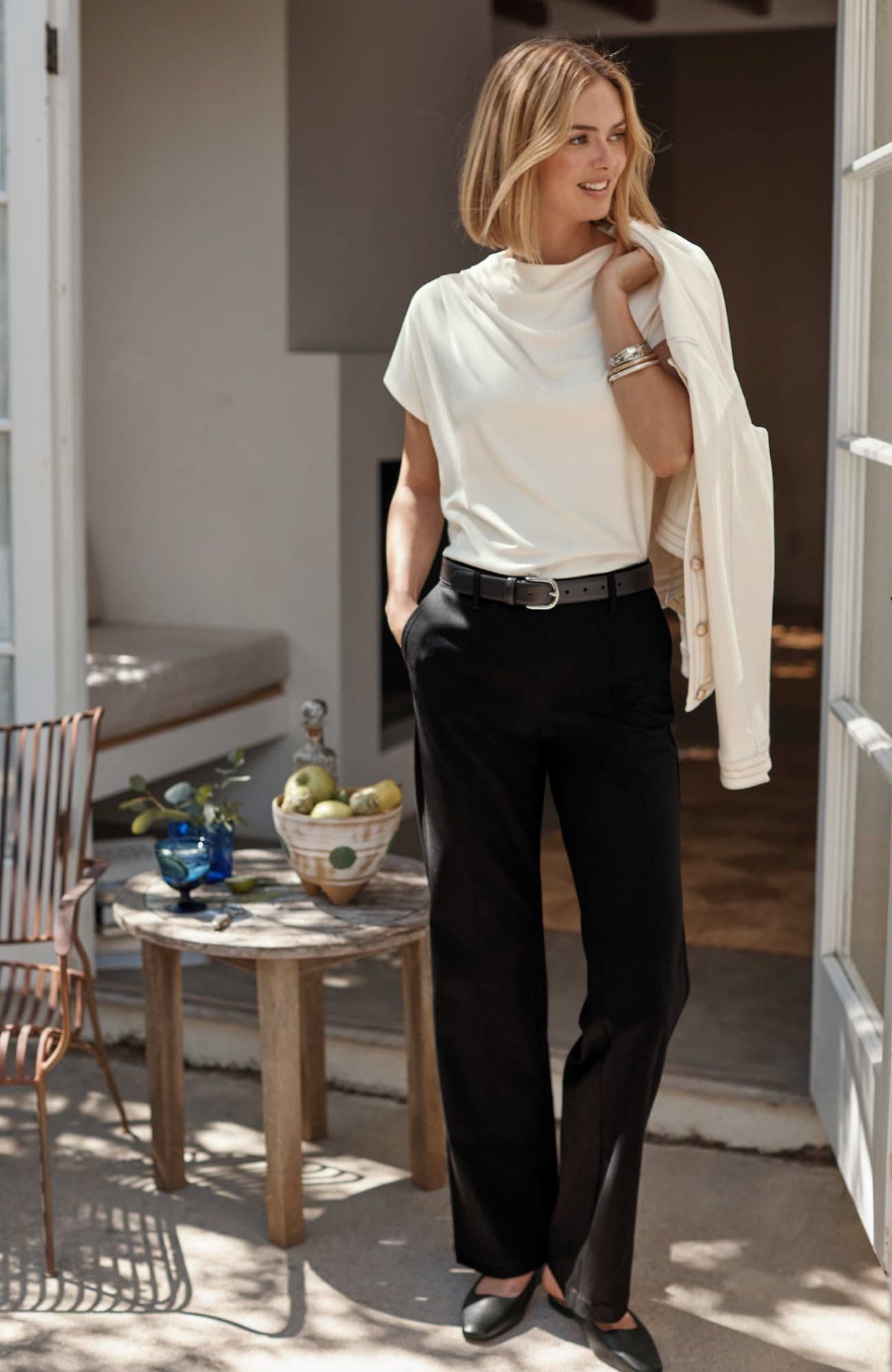 Blonde woman wearing a white knit top, black trousers, and black loafers standing outdoors.