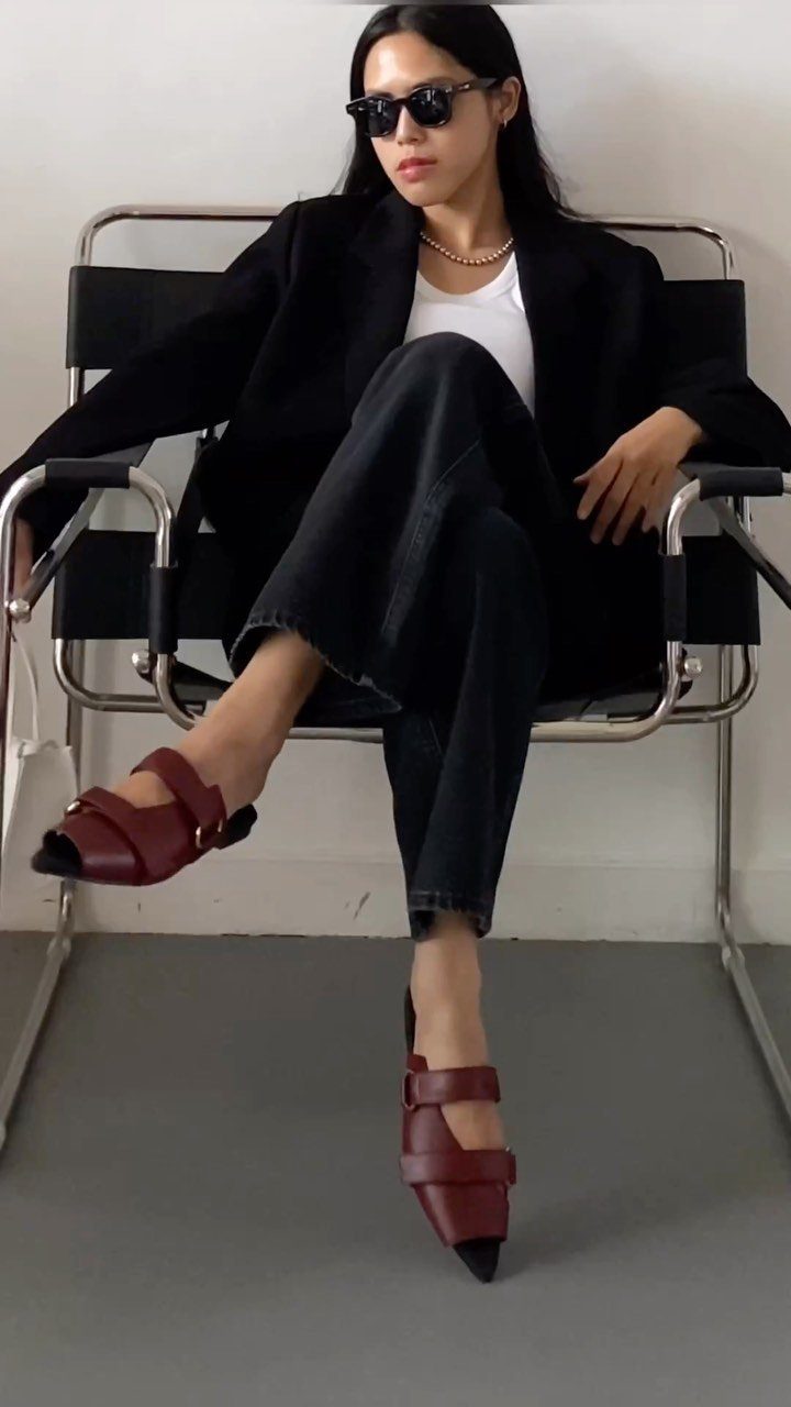 Woman sitting in a chair wearing a black blazer, white top, dark denim, and burgundy sandals.