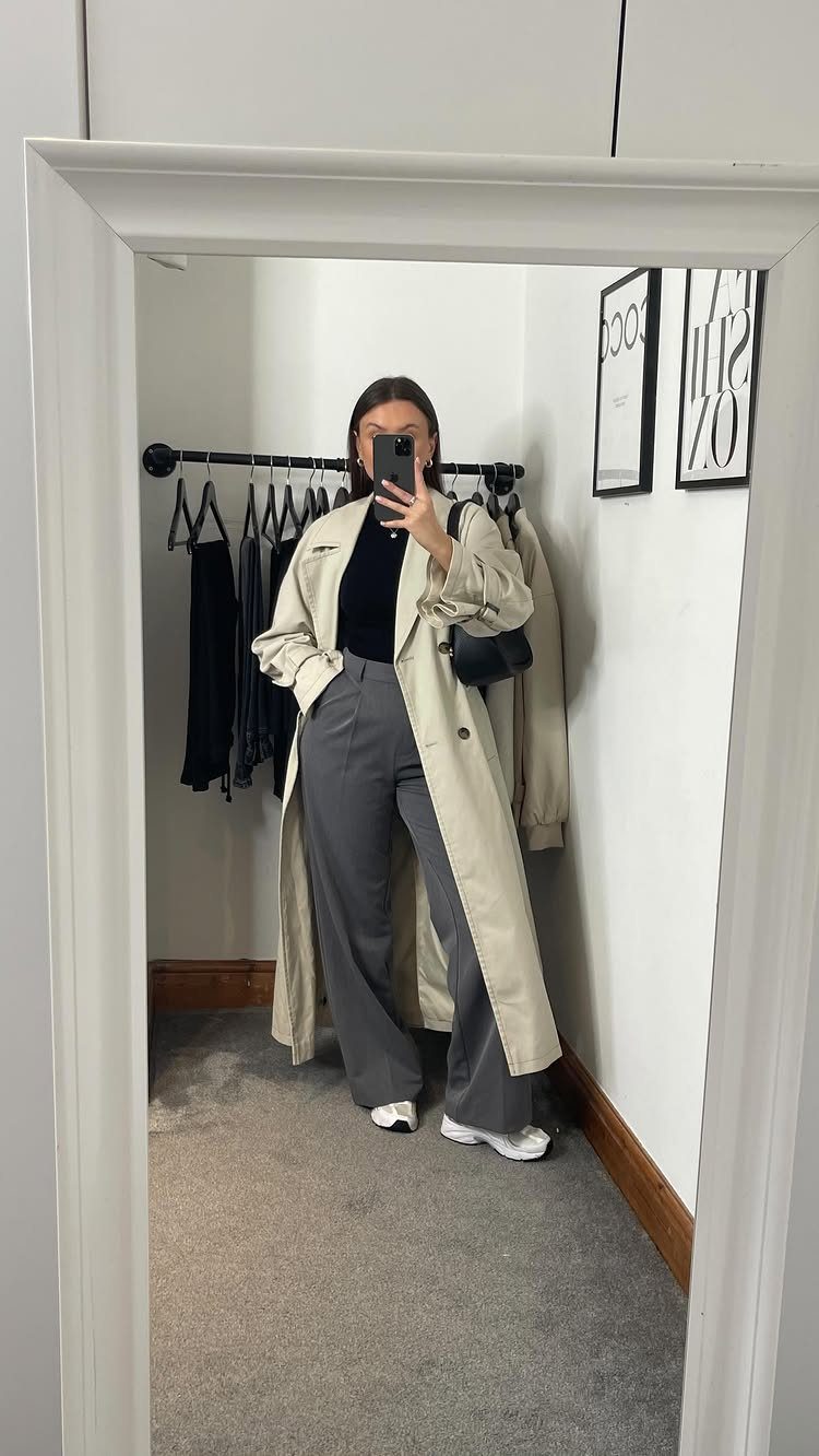Woman in a long beige trench coat, black top, and grey wide-leg trousers posing in a mirror.
