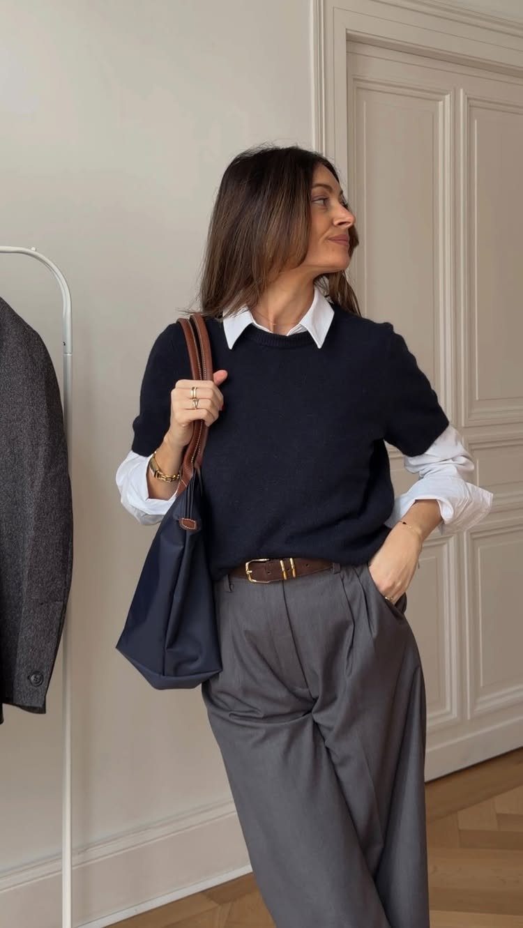 A woman wearing a navy sweater, white collared shirt, and grey trousers while carrying a tote bag.