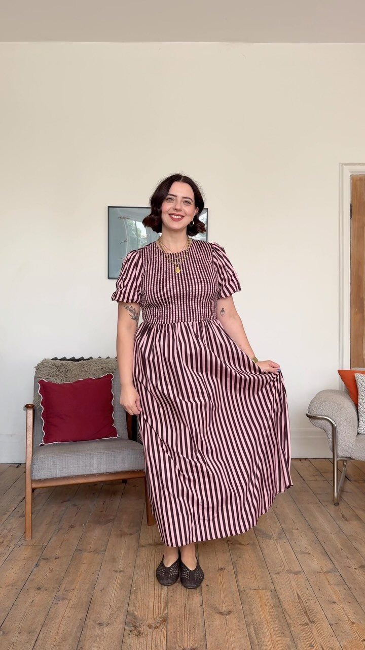 A smiling woman wearing a striped midi dress and dark flats standing in a room.