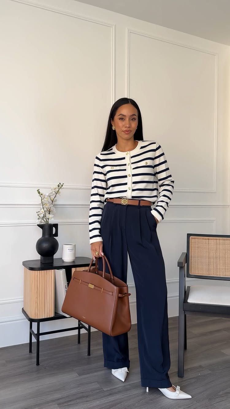 A woman posing in a striped cardigan, navy wide-leg trousers, and white heels holding a brown handbag.