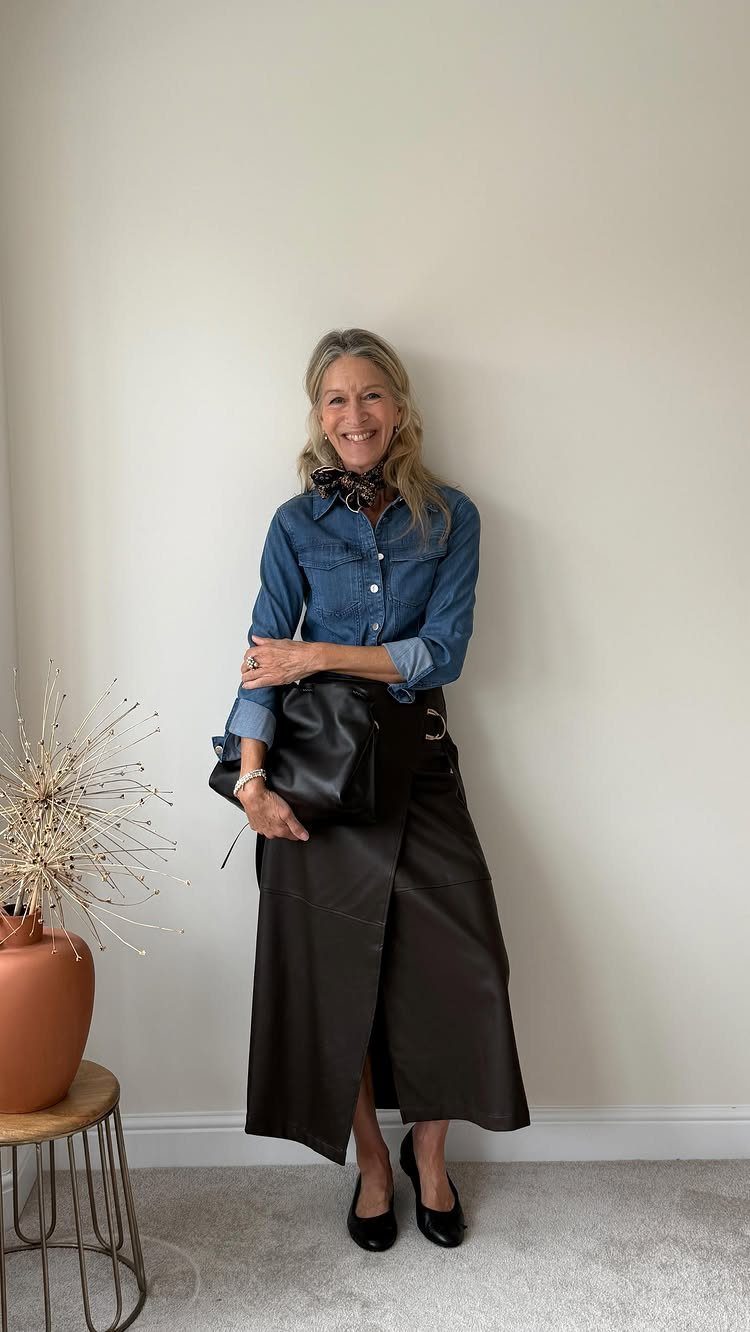 A smiling woman wearing a denim shirt, leather skirt, and black accessories standing against a wall.